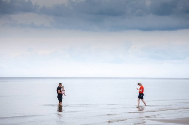 JURMALA, LATVIA - 26 AĞUSTOS 2023: Baltık Denizi kıyısındaki Jurmala 'da anne fotoğraf çekerken bir adam, bir baba, bir çocuk, bir bebek, birbirine bağlanan ve denizin keyfini çıkaran seçici bulanıklık.