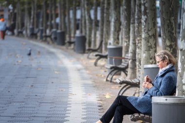 DÜKSELDORF, GERMANY - 7 Kasım 2022: Yaşlı yaşlı bir kadının seçici bulanıklığı, bir bankta oturup yemek yiyor, öğle yemeği yiyor, Almanya 'da alacakaranlıkta,.