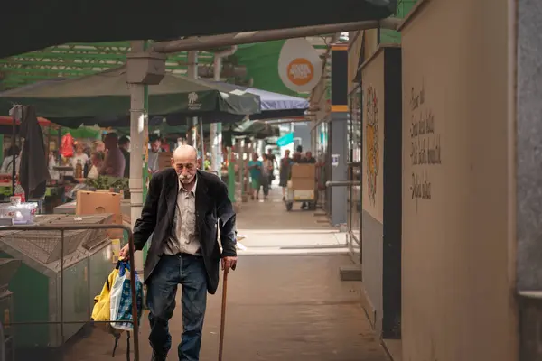 stock image BELGRADE, SERBIA - APRIL 6, 2024: Seective blur on a senior old man with a walking stick passing by the aisles of Belgrade zeleni venac green market with grocery bags.