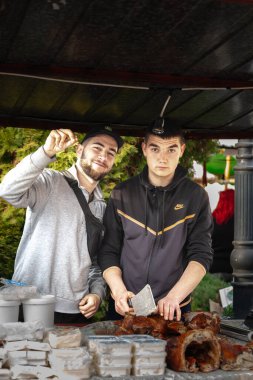 KACAREVO, SERBIA - FEBRUARY 18, 2024: Two Young men, butchers, holding a knife chopping and cutting meat & smiling, in a rural market of Serbia, in Kacarevo, Vojvodina, agricultural province of Serbia. clipart