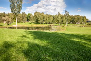 Letonya 'da çayırlarla ve ağaçlarla çevrili açık bir alanda manzaralı bir gölet. Huzurlu doğa sahnesi, manzaranın doğal güzelliğini vurgulayan huzurlu ve resmedilmiş çevreyi yansıtır..