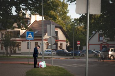 SIGULDA, LATVIA - AUGUST 23, 2023: An elderly man checks his phone while standing next to grocery bags from a supermarket. latvian popuation is ageing. clipart