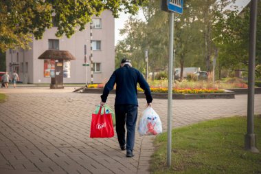 SIGULDA, LATVIA - 23 Ağustos 2023: Sigulda, Letonya 'da bir cadde boyunca birden fazla market poşeti taşıyan yaşlı bir adam yürüyor. Resim, Letonya kentindeki yaşlıların günlük yaşamlarını ve bağımsızlıklarını gösteriyor..