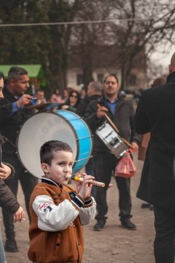 KACAREVO, SERBIA - 18 Şubat 2024: Sırbistan 'ın Kacarevo kentindeki canlı bir pazarda yaşlı müzisyenler arasında flüt çalan genç bir çocuk, kültürel geleneklerin altını çiziyor.