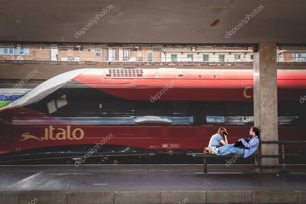 FLORENCE, ITALY - JUNE 15, 2024: Two young people sitting at Florence's Santa Maria Novella station while Italo train passes by in a blur. Italo is a private train carrier in italy, high speed rail.