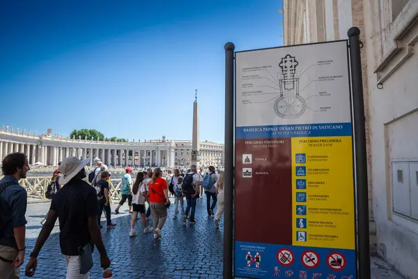 stock image VATICAN - JUNE 15, 2024: Tourists pass by a sign indicating the map and instructions for visitors in Piazza San Pietro (saint peter square), Vatican, with the background of the basilica and surrounding colonnades.