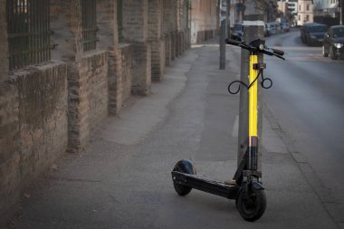 Elektrikli scooter 'daki seçici bulanıklık kaldırımdaki bir direğe güvence altına alınarak çevre dostu ulaşım seçeneklerinin yükselişini ve bunları kentsel altyapıya entegre etmenin zorluklarını temsil ediyor..