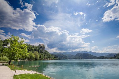 Slovenya 'daki Bled Gölü' nün (Blejsko Jezero) panoramik manzarası, Bled Castle (Blejski Hrad) ile yemyeşil bir uçurum ve güneşli bir gökyüzü altında sakin bir göl. Avrupa 'da popüler bir yaz seyahati merkezi.