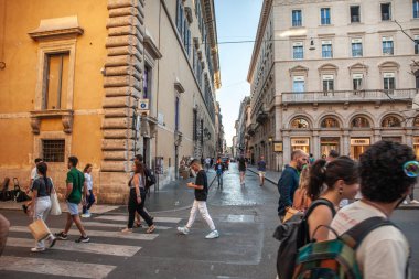 Parlak bir gökyüzü ve yıpranmış duvarcılık altında, insanlar eski Roma 'da sessiz bir cadde boyunca yürüyorlar. Bu şehir tablosu, modern ayak sesleri sanat, politika ve inanç çağlarında yankılandıkça şehirlerin katmanlı tarihini gözler önüne seriyor. Eski taşlar çağdaş taşlarla buluşur