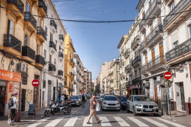 VALENCIA, SPAIN - OCTOBER 13, 2024: A man crosses a pedestrian crossing in the city center on a sunny day, surrounded by historic facades, parked cars, and local businesses reflecting urban life. clipart