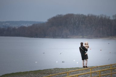 BELGRAD, SERBIA - 12 Ekim 2024: Bir baba Sırbistan 'ın Zemun kentindeki Tuna Nehri' ni izlerken kızını kucağına alıyor. Sahne, sakin bir rıhtım zeminine karşı sessiz bir aile anı yakalıyor..