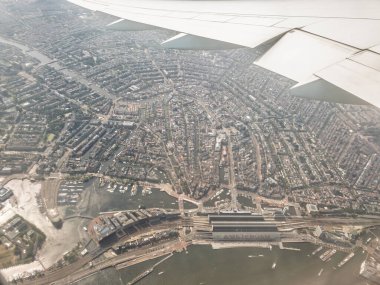 Bir uçak penceresinden Amsterdam, Hollanda 'nın havadan görünüşü, şehir kanalları, tarihi mimari ve Amsterdam Merkez İstasyonu' nun görüntüsü, kentsel yerleşim ve turistik yerleri vurguluyor.