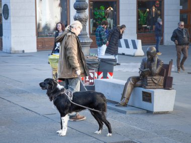 TRIESTE, ITALY - 17 Aralık 2024: İnsanoğlu köpeğini Trieste, İtalya sokaklarında gezdiriyor, bronz bir heykeli gözlemlemek için duruyor, günlük hayatı, evcil hayvan arkadaşlığını ve kentsel kültür sahnesini resmediyor.