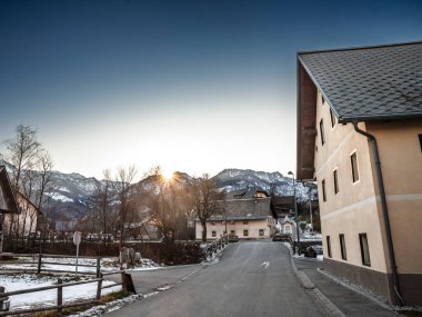 Slovenya 'nın Stara Fuzina kentinde kış alacakaranlığı: Karlı Julian Alpleri' nin arkasında güneş batarken, Alp köylerindeki Pastel çiftlik evleri ve köprülerden sessiz bir yol esiyor