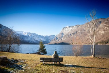 BOHINJ, SLOVENIA - 23 Aralık 2024: Tahta bir bankta oturan yalnız bir yolcu Bohinj Gölü ve karlı Julian Alpleri 'nin sakin sularına bakıyor. Bohinjsko Jezero Slovenya 'nın simgesidir