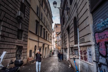 ROME, ITALY - 15 Ocak 2025: Turist, Monti Mahallesi 'ndeki kaldırım taşı caddesinde selfie için bir telefon kaldırırken yayalar ve escooter aksanı tarihi cephelerin dar sokaklarında.