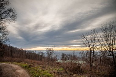 Yapraksız orman ve dolambaçlı toprak bir yol uzanır katmanlı gri bulutların altında sıcak günbatımından oluşan bir şerit ufuk boyunca yanar, sonbaharın sonundaki alacakaranlığın dinginliğini yakar..