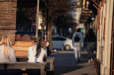 PANCEVO, SERBIA - 3 Mart 2025: Gri kapüşonlu genç bir kadın Pacevo, Sırbistan 'da bir kafe bankında oturuyor, elinde yanan bir sigara, akşam ışığında süzülen bir duman, yoldan geçenler arka planda bulanıklaşırken.