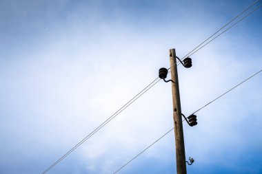 Sırbistan 'da soluk mavi kırsal bir gökyüzü boyunca çaprazlama uzanan kahverengi seramik yalıtkan ve ikiz elektrik hatlarıyla donatılmış yaşlanmış ahşap bir elektrik direğinin minimalist manzarası.
