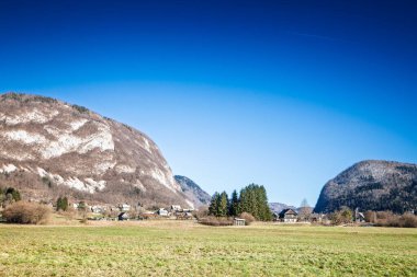 Geniş Alp çayırı, Slovenya 'nın Bohinj Vadisi' ndeki Stara Fuzina kümelenmiş evleri, orman kireçtaşı yamaçları ve Triglav ulusal parkında kusursuz bir kış ortasında mavi gökyüzü ile çevrelenmiş durumda..