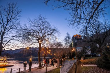 BLED, SLOVENIA - 17 ARALIK 2024: Slovenya Gölü üzerindeki Noel süsleri Ziyaretçilerin, şenlikli kıyı ışıklarının fotoğrafını çekerken Bled; uçurumun tepesindeki Bled kalesi, kışın sakin bir akşamda suyun üzerinde parlıyor