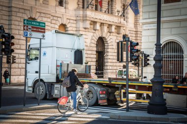 TRIESTE, ITALY - 17 Aralık 2024: İtalya 'daki Trieste City Center' daki kalabalık Piazza della Liberta 'da uzun ve eklemli bir kamyon kükreyerek geçerken Bits marka bir bisikletin üzerinde sırt çantalı bir bisikletçi zebra şeritlerinde bekliyor