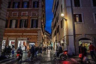 ROMA, İTALYA - 15 OCAK 2025: Tarihsel merkezde akşam sokak manzarası alışveriş yapanların ve yerlilerin parke bir caddede yürüdüğü aydınlanmış butikler, scooterlar ve asırlık cephelerle dolu.