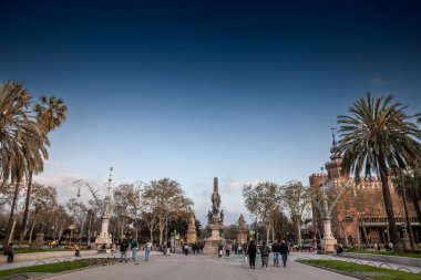 BARCELONA, SPAIN - 12 Nisan 2025: İnsanlar Passeig de Lluis Şirketleri boyunca palmiyeler ve kırmızı tuğlalı Castell Tres Ejderhaları tarafından çerçevelenmiş Rius i Taulet Anıtı 'na doğru yürürler..