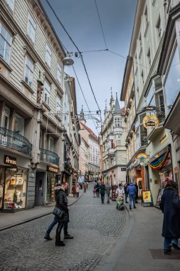 GRAZ, AUSTRIA - 17 Aralık 2024: Pedestrianlar Graz Innere Stadt 'taki kaldırımlı Sporgasse' daki dükkanlara bakıyorlar; renkli tarihi cepheler bulutlu bir kış öğleden sonrasında hafif eğimli şeridi çerçeveliyorlar.