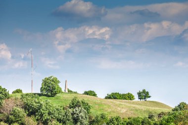 Sırbistan 'ın Voyvodina kentindeki Zagajicka brda Panoraması: çimenli kum tepeleri, dağınık meşe ağaçları, ince bir rüzgar direği ve zagajicka brda obelisk yay bulutlarıyla kaplı geniş bir gökyüzüne karşı yükseliyor.