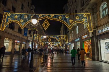 BELGRAD, SERBIA - 12 Aralık 2024: Akşam alışverişçileri butikler ve taş döşemeler arasında yürürken Belgrad Kneza Mihailova yaya yolu üzerinde Noel aydınlatmaları yansıtıyor