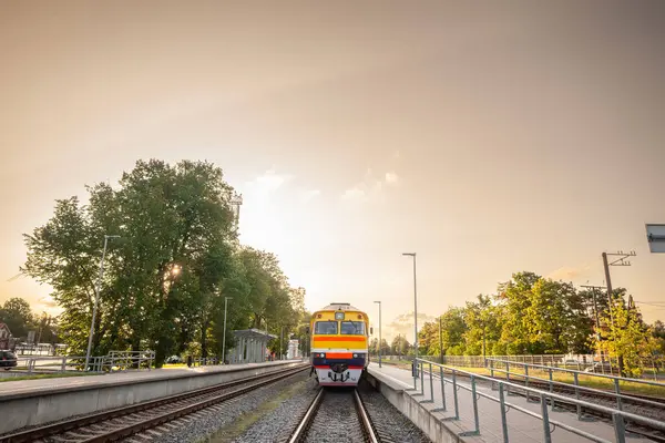 Letonya Demiryolları DMU, Letonya 'daki Sigulda istasyonunda altın gece ışığı gökyüzünü doldururken bekliyor; sarı tren, boş platformlar ve kalkışa hazır, yaklaşan raylar arasında duruyor..
