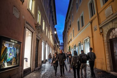 ROME, ITALY - 15 OCAK 2025: Eski Roma 'da dar bir kaldırım taşı caddesi boyunca alışveriş yapan yayalar, parlayan mağazalar, tarihi cepheler ve lamba ışıklı pencerelerle çevrili.