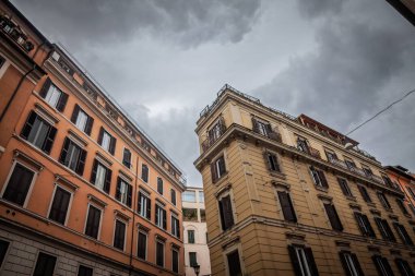 Ochre ve sarı 19. yüzyıla ait apartman blokları, Roma 'nın tarihi merkezindeki bulutlu gökyüzünün altında ahşap kepenkler, duvarcılık ayrıntıları ve dar yerleşim yoğunluğu vurgulanıyor..