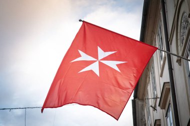 Malta 'nın Egemen Askeri Düzeni' nin kırmızı bayrağı. Beyaz sekiz köşeli Malta haçı, Ljubljana 'da klasik bir cephenin yanında soluk bir gökyüzüne doğru dalgalanır. Şövalyelik Nişanı' nın kalıcı amblemini vurgular..