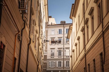İtalya 'da güneşli bir avluya doğru Trieste' nin tarihi çekirdeğindeki dar bir sokağa bakan duvarlar, kepenkli pencereler arasına asılmış bir çamaşır ipi örer ve İtalyan Akdeniz mimarisini yakalar..