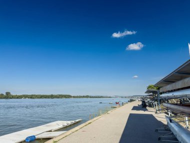 BELGRAD, SERBIA - 22 Nisan 2025: Sırbistan 'ın Zemun kentindeki Zemunski kej' de nehir kenarındaki geniş gezinti alanı, sakin Tuna suları ve açık mavi bir yaz gökyüzü yanında barınakların altında kürek kabukları depolanmış durumda.