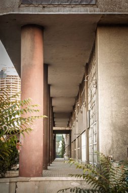 Düşük açı perspektifi aşağı uzun gölgeli bir açık koridor aşağı doğru geniş kırmızı silindirik kolonlar sol ve sıra sıra endüstriyel duvar lambaları ve metal kapılar sağ, derin bir çizgisel kaybolma noktası oluşturur..