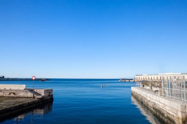 İtalya, Trieste liman rıhtımlarından Adriyatik Denizi 'nin parlak sabah manzarası, sakin mavi suları, kanaldaki şamandıraları, mavi gökyüzünün altındaki uzak pilot römorkörleri ve rıhtım depolarını gösteriyor..