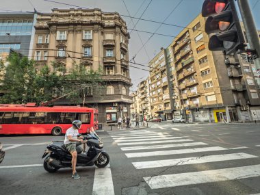 BELGRAD, SERBIA - 6 Haziran 2025: Sırbistan 'ın orta kesimindeki yoğun bir kavşakta, ön cephelerin çerçevelediği kırmızı bir tramvayın yanında kırmızı ışıkta bekleyen scooter sürücüsü.
