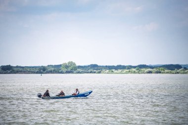 RAM, SERBIA - 13 Mayıs 2025: Sırbistan 'ın Ram kenti yakınlarındaki Tuna Nehri' nde üç balıkçı taşıyan mavi bir sürat teknesi süzülerek geniş nehir üzerinde hafif bir iz bırakırken, yeşil nehir kıyısı ormanları uzak kıyı şeridi boyunca uzanır.