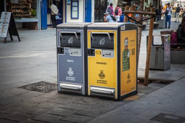 VALENCIA, İspanya - 15 Nisan 2025: Valencia, İspanya 'da bir yaya caddesinde geri dönüşüm ve atık konteynırları. Sarı kabartmalar ve gri kabartmalar Ajuntament de Valencia logosunu gösteriyor.
