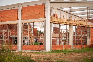 Beton kolonlar, tuğla döşemeler, iskeleler ve çelik kirişlerle büyük bir alana yeni bir depo inşa ediliyor. Çerçeve, gelecekteki bir lojistik tesisi için devam eden bir yapıya işaret koyuyor..