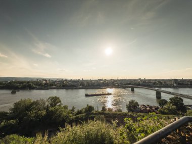 Sırbistan 'ın kuzeyinde Varadinski köprüsü veya duga ile Tuna Nehri üzerinden Novi Sad' a doğru yüksek bir bakış açısı. Öğleden sonra güneşi nehirde parlıyor, önplanda yeşillik kentsel bir manzarayı çerçeveliyor.