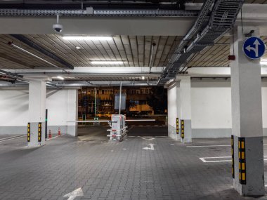 Bariyer, bilet makinesi ve floresan ışık altında CCTV ile bir yeraltı otoparkında gece görüşü. Boş sokaklar, oklar ve beton sütunlar kontrollü erişimi olan bir sokak çıkışına çıkıyor..