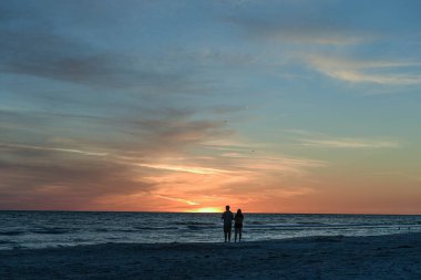 Mavi gökyüzü, martılar, kıyıda çift, ufukta güneş.