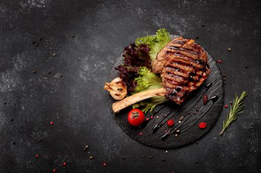 Kemiklerde Tomahawk bifteği, otlar, domatesler ve sarımsak, siyah tahtada, koyu betonda