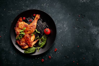 Kızarmış tavuğun yarısı, siyah bir tabakta, otlar ve domateslerle, koyu bir betonun üzerinde.