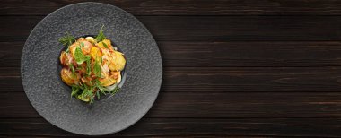 Fried potatoes with garlic and herbs on a black plate, on a wooden background. Banner