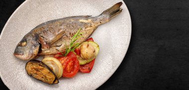 Fried dorado fish, in a plate, with grilled vegetables, on a dark concrete background. Banner